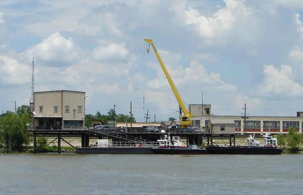 A yellow crane on a pier lifts cargo next to a small building, with two tugboats docked alongside on the river.