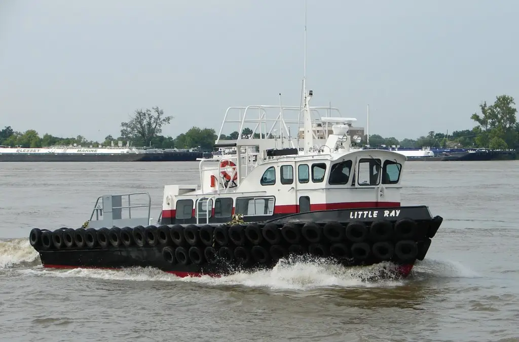 A small tugboat named "Little Ray" moves along a river, surrounded by several large black tires on its sides, with trees and barges visible in the background.