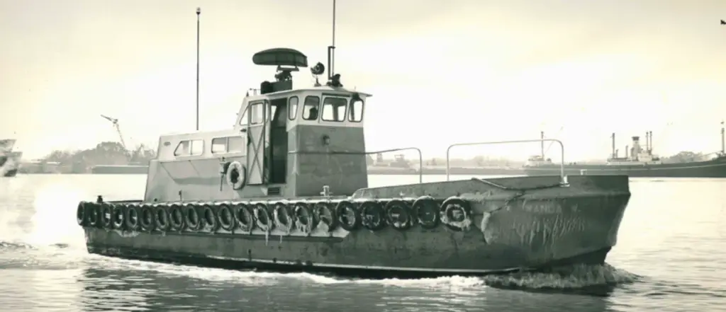 A small tugboat with rubber tires along its sides moves through calm water; industrial ships and cranes are visible in the background.