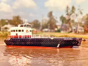 A small tugboat named “Miss Rae Anne,” part of the Original Fleet, floats on a river, with black tires lining its sides and trees and buildings visible in the background.