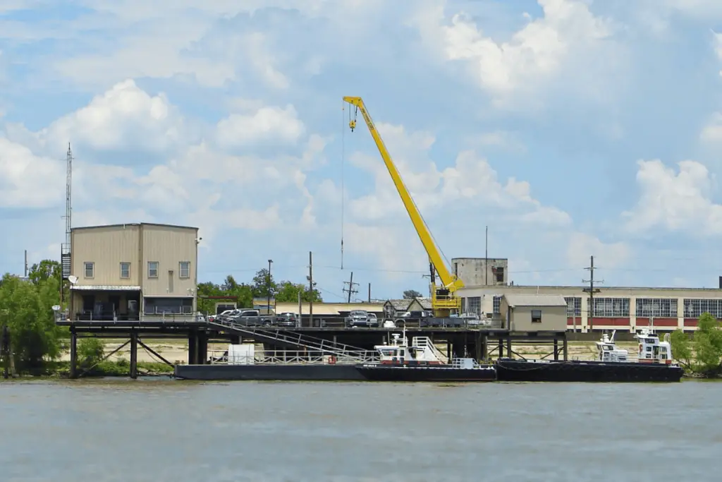 A yellow crane stands on a riverside dock with two boats moored alongside; industrial buildings and trees are visible in the background under a partly cloudy sky.