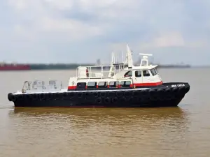 A black and white utility boat from the Original Fleet, with rubber fenders and the name "Miss Caitlin" on the bow, floats on calm brown water under a cloudy sky.