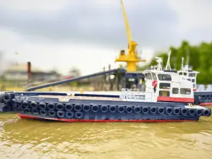 An Original Fleet tugboat with tires along its sides floats on brown water near a port, framed by cranes and industrial structures in the background.