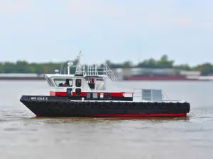 A black, red, and white utility boat named "Miss Leslie II" from the Original Fleet travels on a river, with trees and blurred buildings visible in the background.