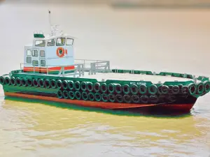 A small cargo boat named "MCS ARIHAN" from the Original Fleet floats on murky water, with rows of black tires attached to its sides as fenders.