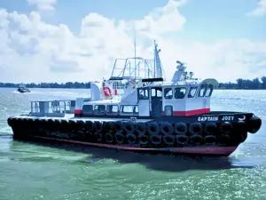 A black and white tugboat named "Captain Joey" floats on the water, surrounded by tires along its sides, with another boat visible in the background.